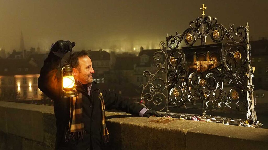 Mateo, guía oficial en Praga, con un farolillo encendido junto a la reja de San Juan Nepomuceno en el Puente de Carlos bajo la niebla nocturna.