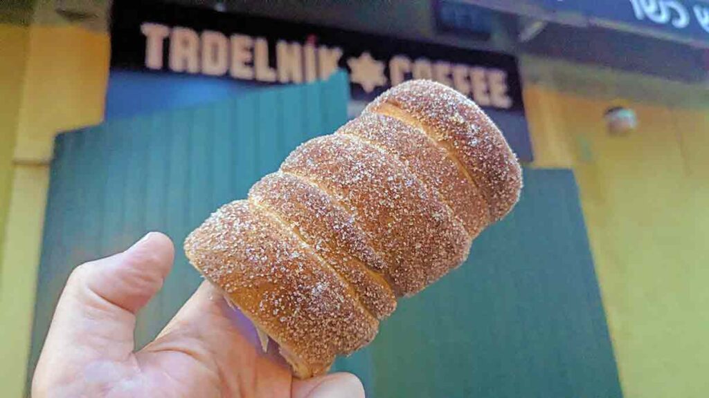 Primer plano de un dulce trdelník cubierto de azúcar y canela sostenido con la mano frente a un puesto del barrio judío de Praga.